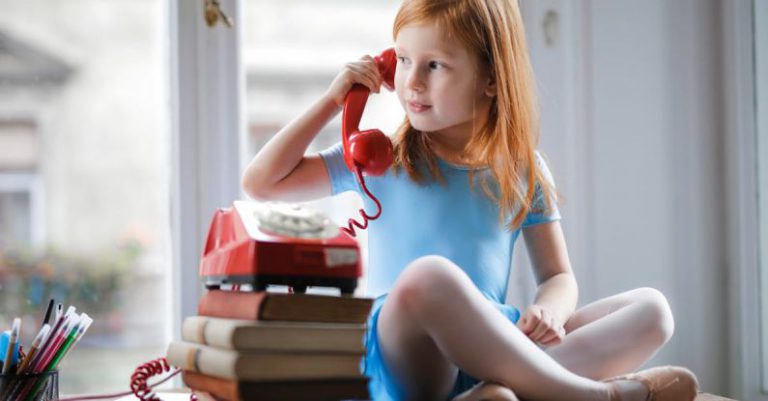 Small Talk - Low angle of calm redhead preteen lady in blue dress and beige sandals looking away and having phone call using retro disk telephone on stack of books while sitting with legs crossed on wooden table against window at home