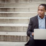Startup Steps - Positive young black businessman using laptop on stairs