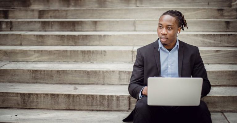 Startup Steps - Positive young black businessman using laptop on stairs