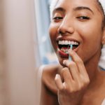Morning Habits - Smiling African American female with white towel on head cleaning teeth with dental flosser in bathroom