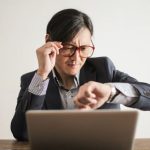Entrepreneur Time - Young frowning man in suit and glasses looking at wristwatch while waiting for appointment sitting at desk with laptop
