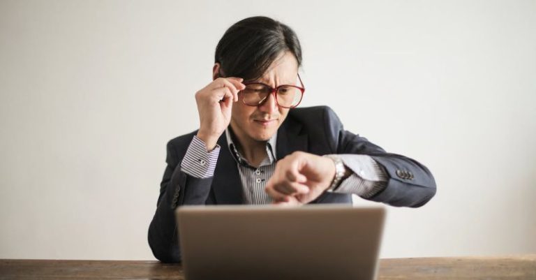 Entrepreneur Time - Young frowning man in suit and glasses looking at wristwatch while waiting for appointment sitting at desk with laptop