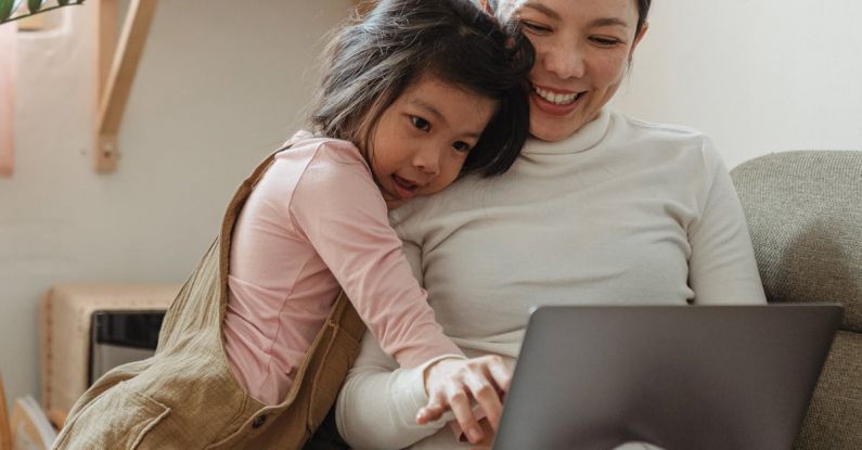 Parent Work Balance - Cheerful smiling Asian woman browsing modern netbook while hugging with cute content daughter on comfy sofa in cozy living room