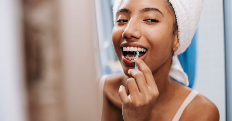 Morning Habits - Smiling African American female with white towel on head cleaning teeth with dental flosser in bathroom