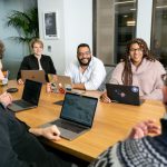 Leadership - four people all on laptops, two men and two women, listen to person talking in a board meeting