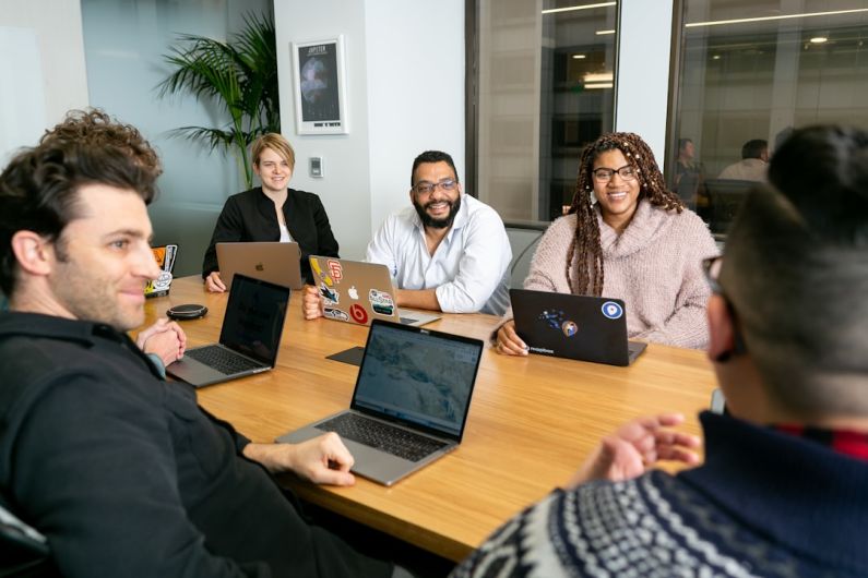 Leadership - four people all on laptops, two men and two women, listen to person talking in a board meeting